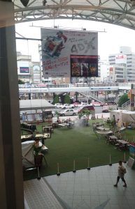 An area of fake grass with food stalls and chairs/tables on it, between a railway station and a busy road & bus station