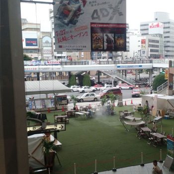 An area of fake grass with food stalls and chairs/tables on it, between a railway station and a busy road & bus station