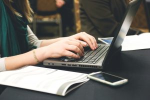 Closeup of a woman typing on a laptop with a notebook and a mobile phone next to her.