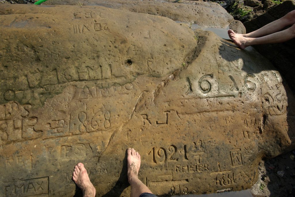 An exposed riverbed rock with carvings of dates visible back to 1616. Some people are resting their bare feet against it.